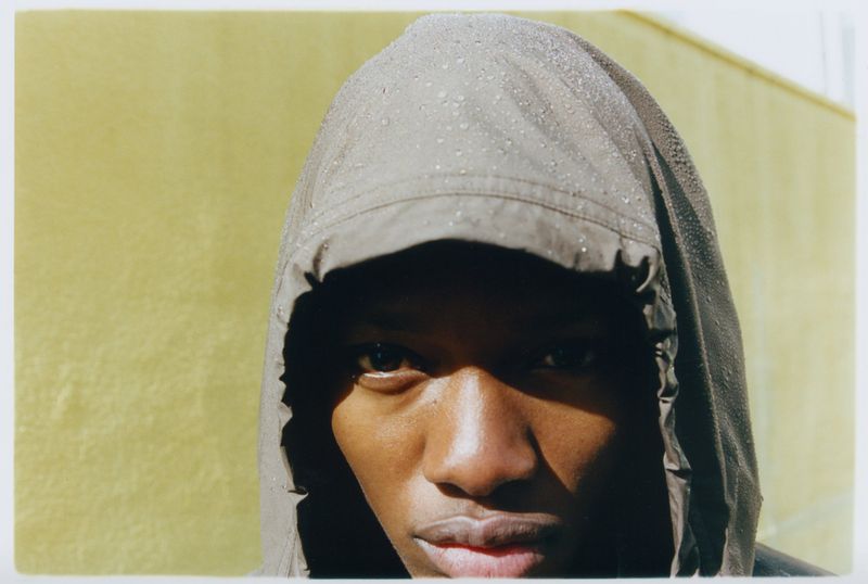 Close-up portrait of a Black adult male wearing a hooded grey jacket with water droplets, set against a yellow outdoor wall.