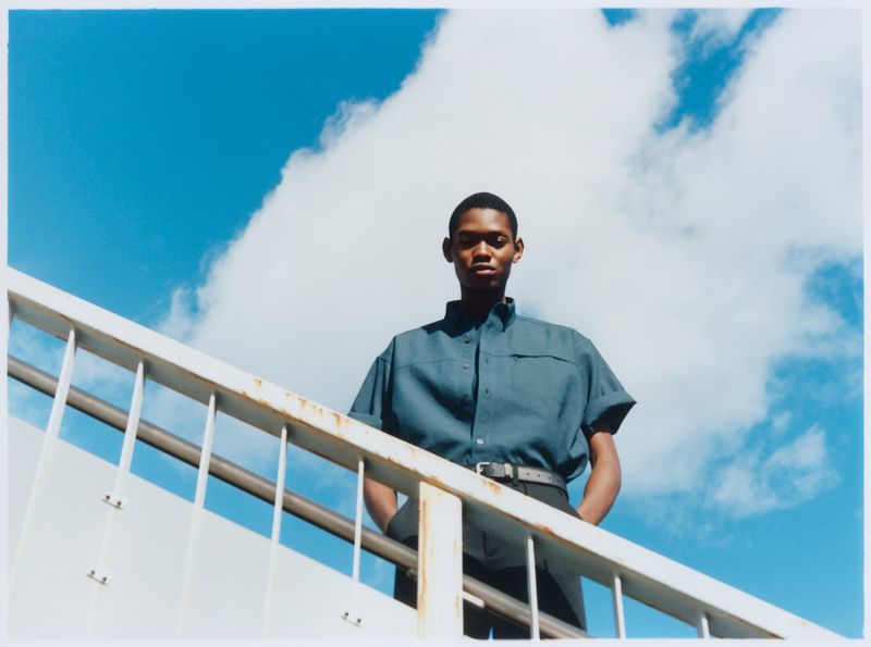 Portrait of a young African-American man in a blue shirt, standing on stairs with a white railing against a bright blue sky and clouds.