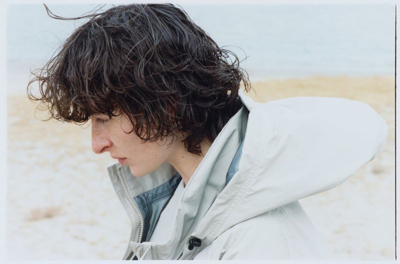 Close-up shot of a young adult with dark, wavy hair wearing a hooded jacket on a beach. The sky and water blend in the background.