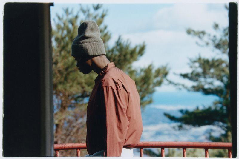 Side profile of a young Black man wearing a beanie and orange shirt looking down, with trees and sky in the background.