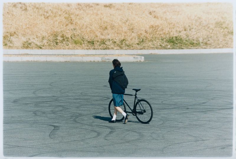 Adult walks with black track bicycle across asphalt in open space with dried grass and bright sun, wearing backpack and comfortable clothes.