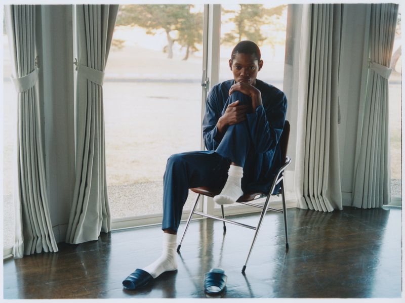 An adult African-American man sits cross-legged on a chair in blue clothing, white socks, and sandals, gazing thoughtfully in a well-lit room.