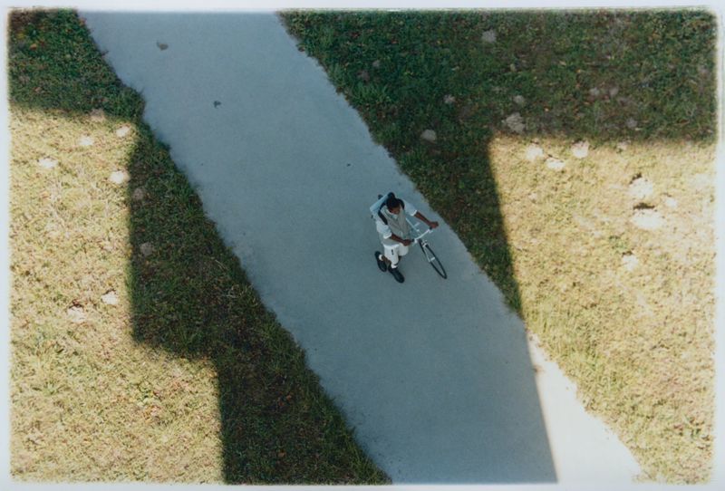 Top-down view of an adult riding a tricycle on a concrete path, surrounded by green grass under natural daylight, creating shadows.
