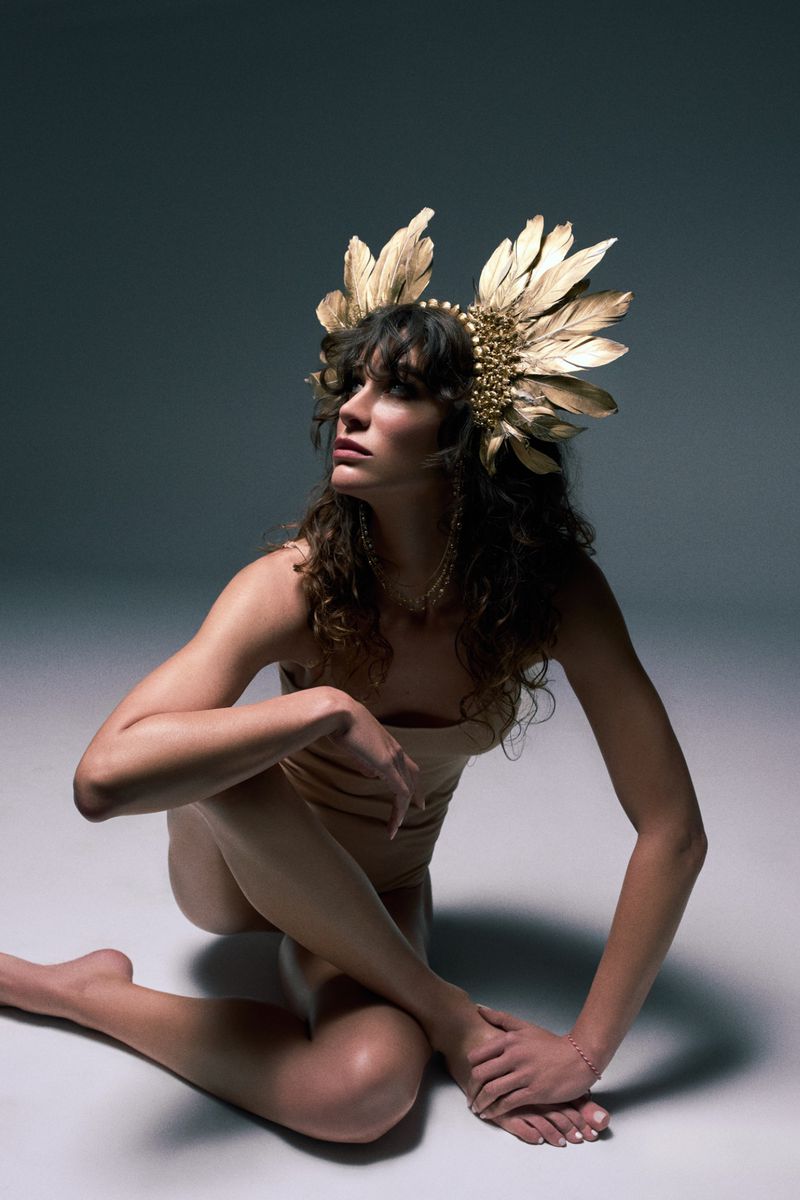 Woman in tan bodysuit and gold feathered headdress poses barefoot in a photo studio. Grey gradient background, medium shot, portrait orientation.