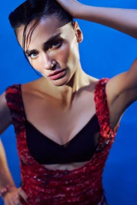 Close-up portrait of a woman with wet-look dark hair wearing a red patterned top against a vibrant blue studio background.