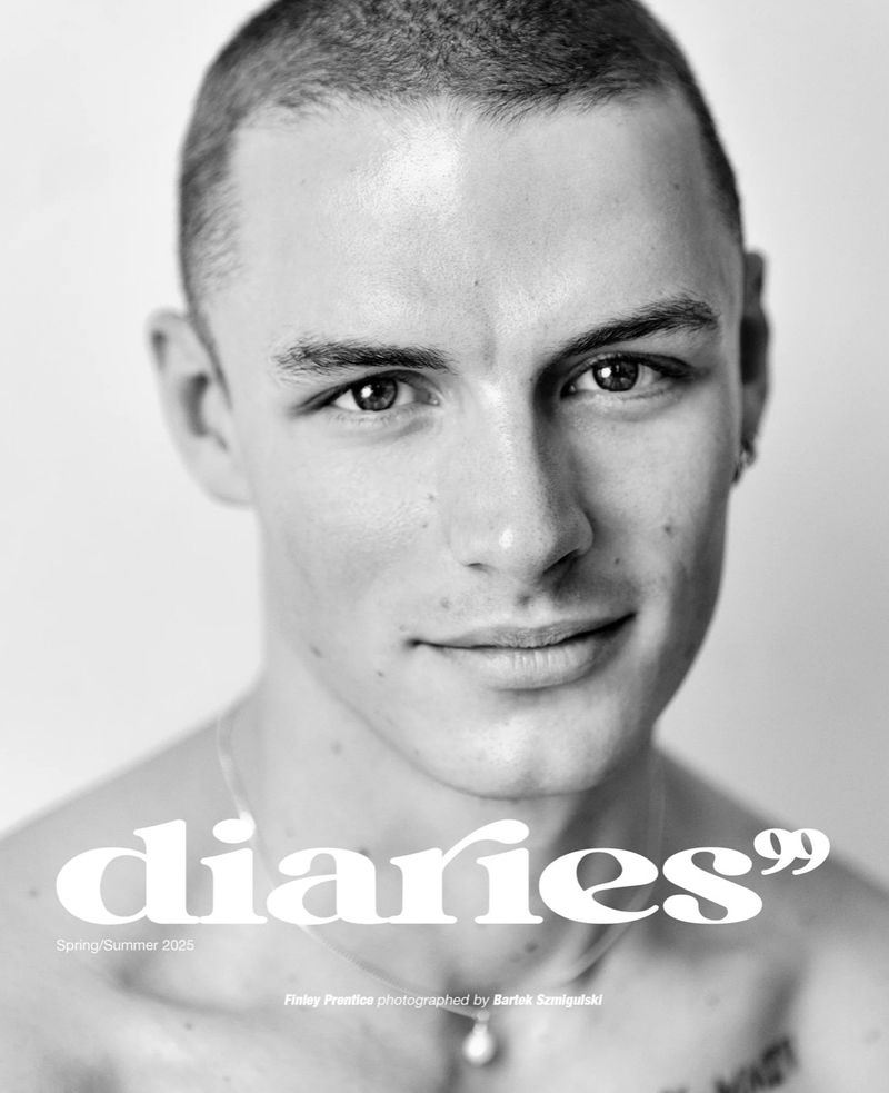 Black-and-white portrait of a handsome young man with a buzzcut, wearing a pearl necklace, against a clean, light-grey studio backdrop.