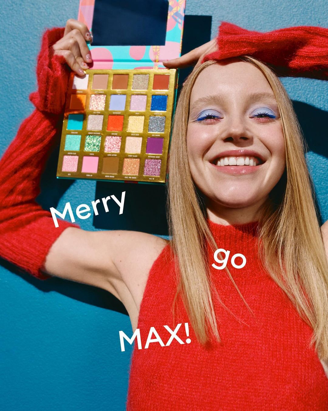 Smiling woman holds a colorful eyeshadow palette, showcasing various shades against a bright blue background in an indoor setting.