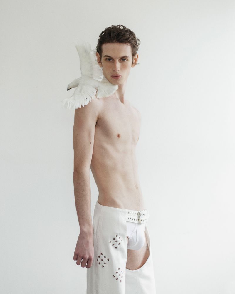 Male model poses topless in chaps with a white dove perched on his shoulder against a white background in a fashion editorial.