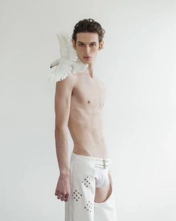 Male model poses topless in chaps with a white dove perched on his shoulder against a white background in a fashion editorial.