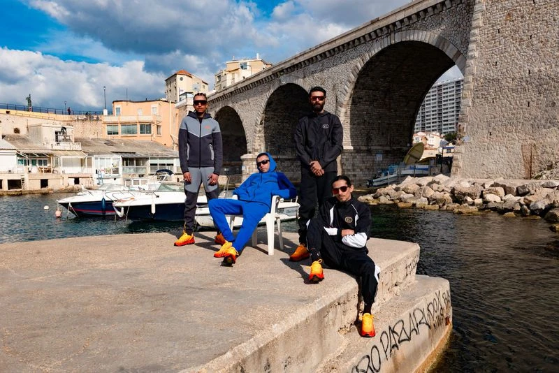 A group of four stylish young men pose on a concrete pier in front of a stone bridge and a blue harbor with boats and distant buildings.