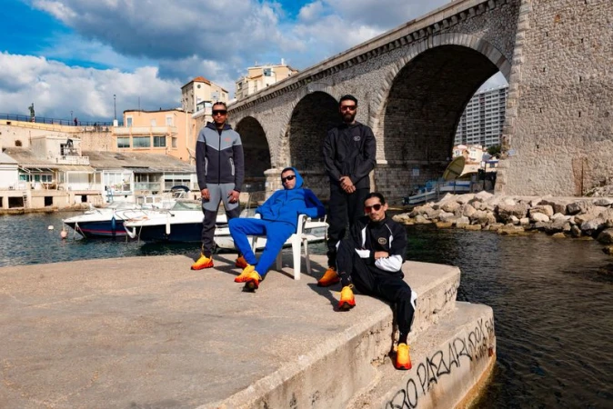 A group of four stylish young men pose on a concrete pier in front of a stone bridge and a blue harbor with boats and distant buildings.