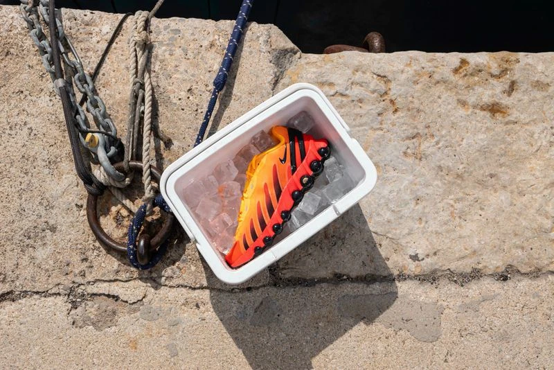 Orange and black athletic shoe sits in a white container with ice cubes, placed on a concrete surface with ropes and metal rings.
