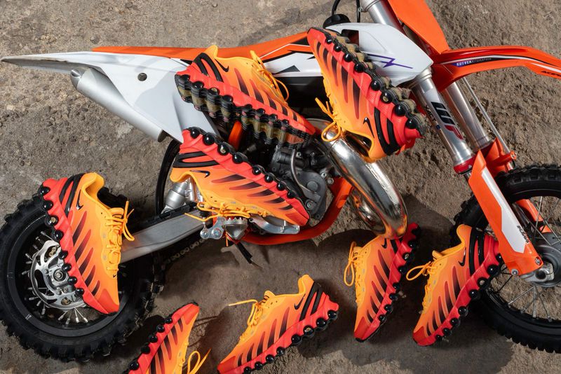Orange and black running shoes are arranged around a parked orange and white motocross bike on a dirt surface, in a product shot.