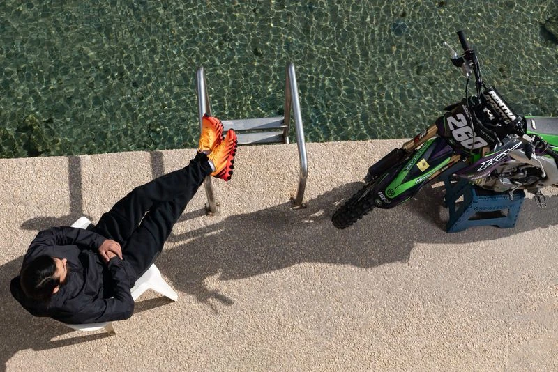 Person relaxes by pool next to motocross bike. Sunlight casts shadows on the tiled surface and turquoise water in outdoor setting.