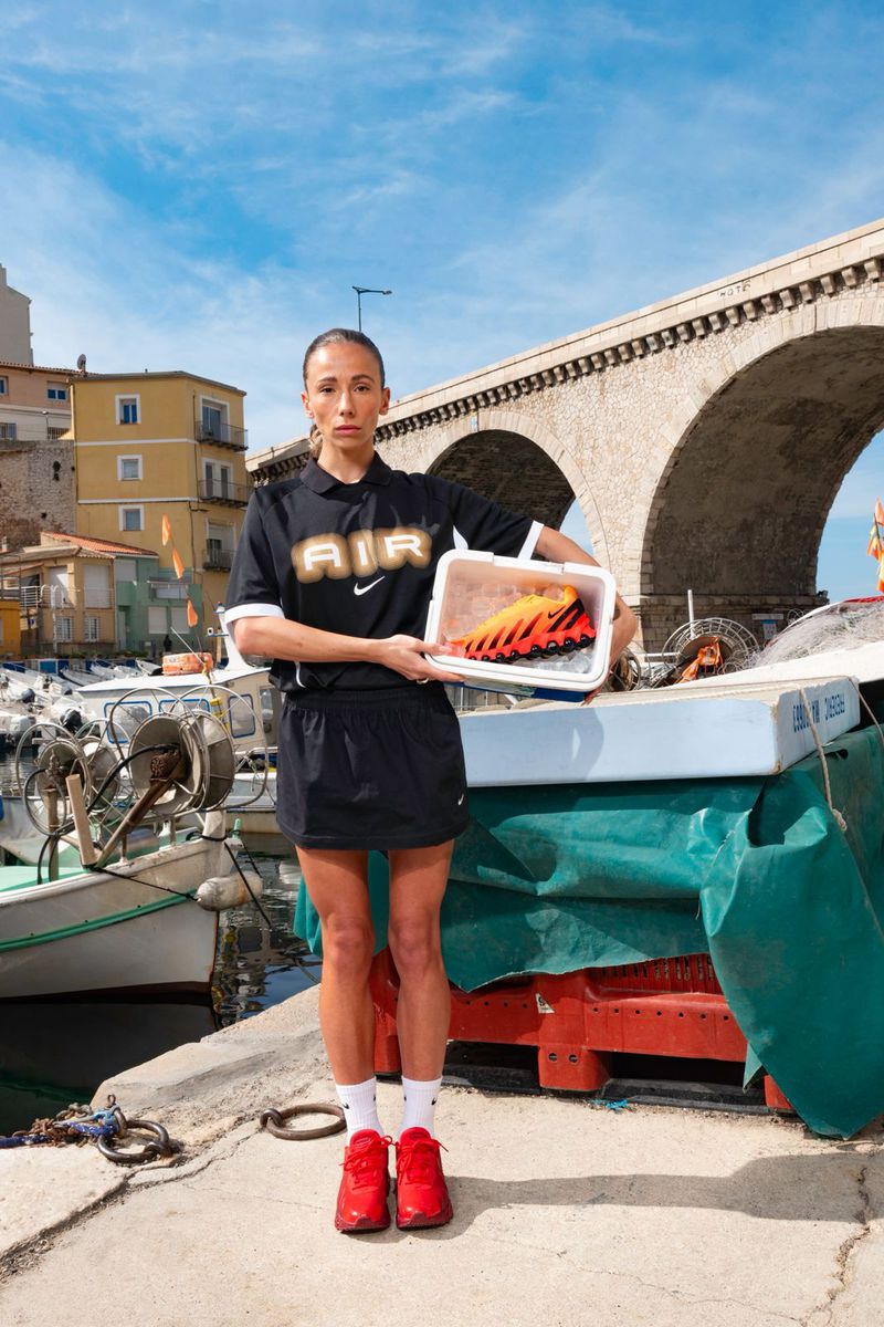 Woman in black sportswear holds orange sneakers in a container outdoors against a backdrop of boats, harbor and an arched bridge.
