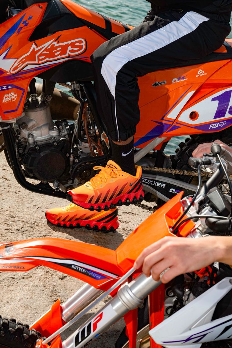 Person in black and white track pants sits on an orange and white motocross bike, with matching orange shoes on a sandy surface.
