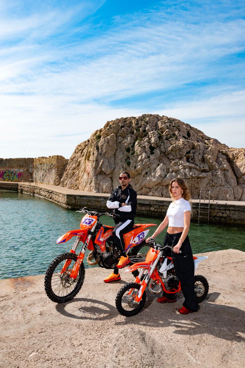 Two people, adult male and female, pose with two orange KTM dirt bikes on a concrete platform near a waterfront on a sunny day.