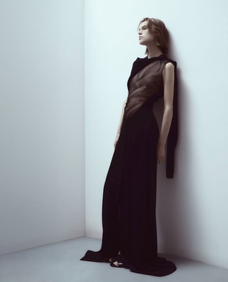 Woman in a black evening gown leans against a corner wall. She gazes upward in a studio setting with neutral, diffused lighting.