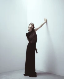 Model wearing a black dress leaning against a white corner wall in a studio setting