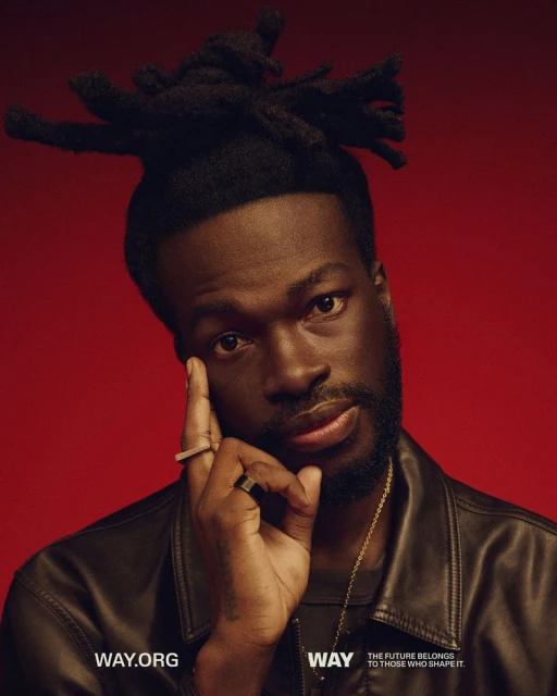 Close-up portrait of a Black man with dreadlocks wearing a leather jacket against a red backdrop, showcasing a ring and gold chain.