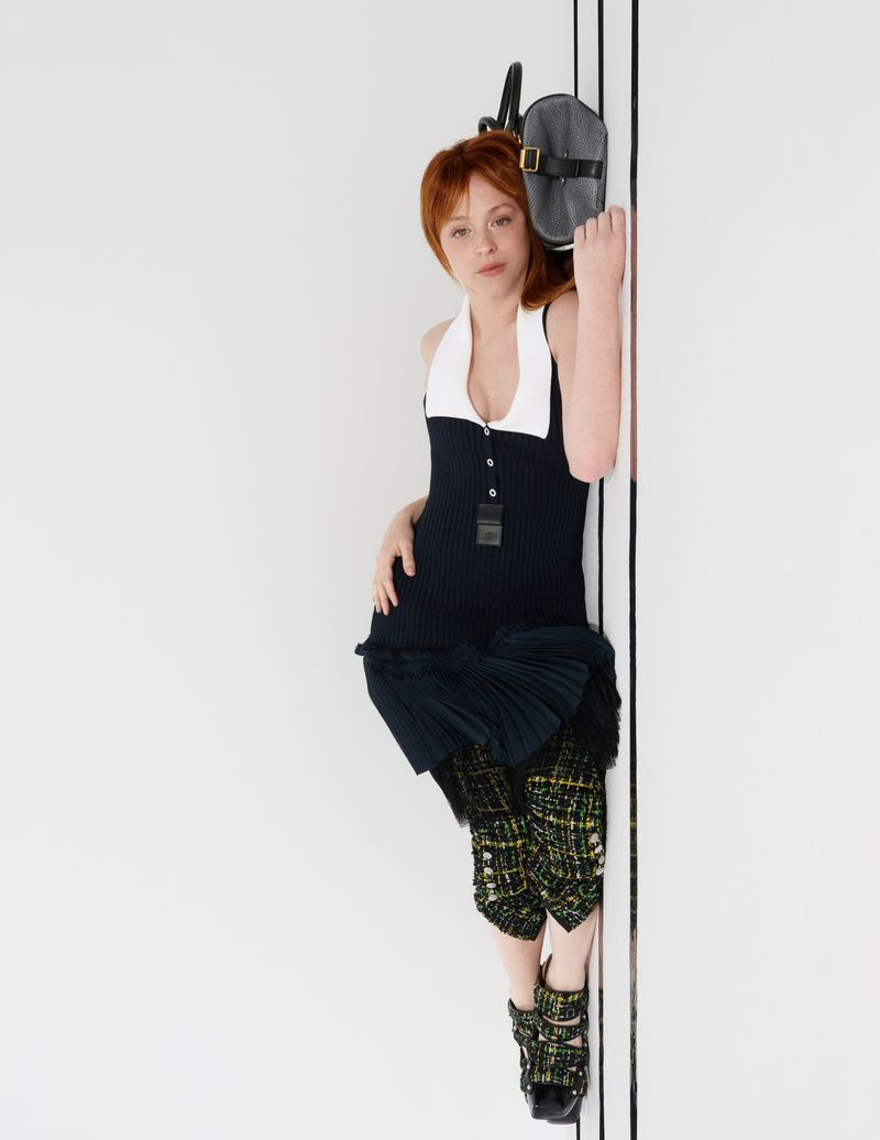 Red-haired woman poses in a sleeveless navy dress with white collar, patterned cropped trousers, and buckled flats against a white wall.