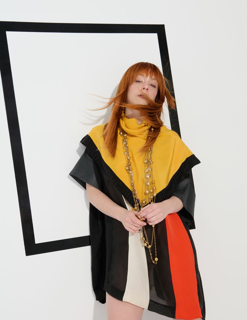 A woman poses wearing a colourful dress and long beaded necklaces, next to a black rectangle on a white background in a fashion shot.