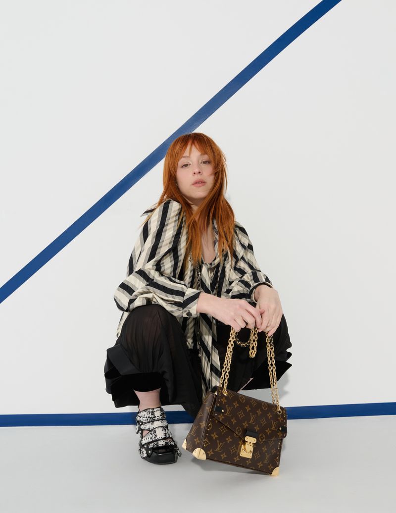 A woman with red hair sits and holds a brown Louis Vuitton purse with gold hardware, wearing a striped shirt and black pants.