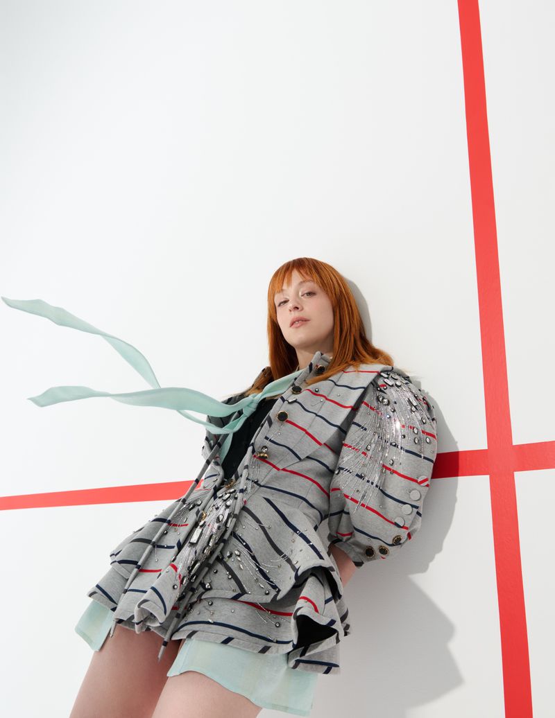 Red-haired woman in a grey striped jacket and shorts posing in front of a white wall with red lines, fashion forward and bold.