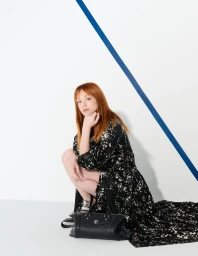 Red-haired woman in a black patterned dress squatting next to a black handbag against a white background with a blue stripe