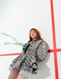 Model with red hair wearing a grey striped jacket leaning against a white wall with red stripes for Marie Claire Spain