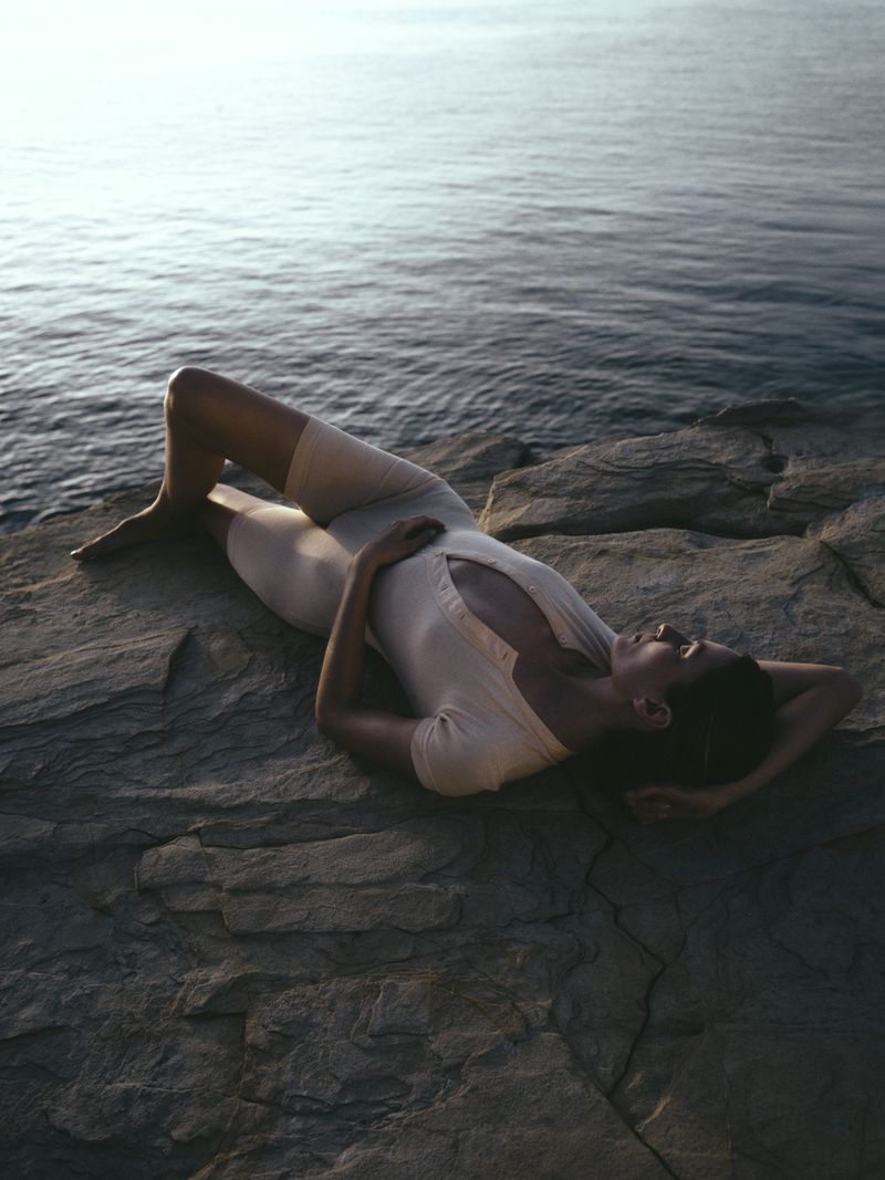 Woman in swimsuit lying on rocky shore, ocean backdrop. Relaxed pose, arms behind her head. Serene, natural light and seascape.
