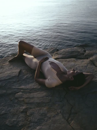 Woman in swimsuit lying on rocky shore, ocean backdrop. Relaxed pose, arms behind her head. Serene, natural light and seascape.