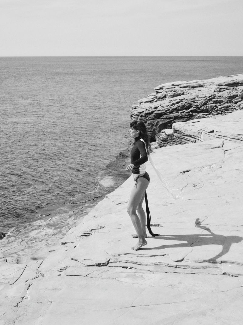 Monochrome shot of a woman in a swimsuit standing on a rocky cliff overlooking the ocean on a sunny day, creating a striking coastal scene.