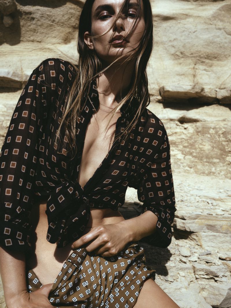Adult female model in outdoor setting wearing patterned blouse and skirt, posing against a weathered rock background, fashion photography.