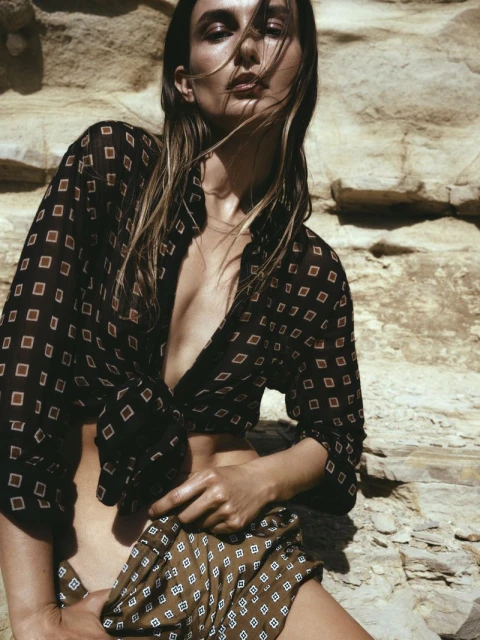 Adult female model in outdoor setting wearing patterned blouse and skirt, posing against a weathered rock background, fashion photography.