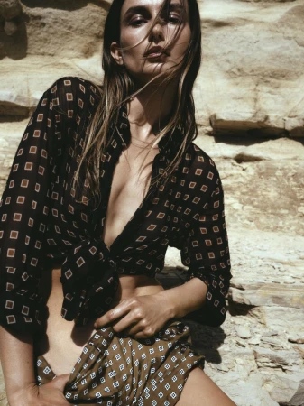 Adult female model in outdoor setting wearing patterned blouse and skirt, posing against a weathered rock background, fashion photography.