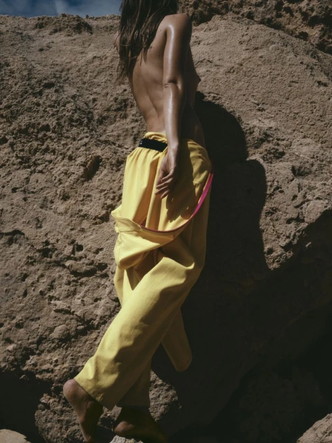 Back view of adult woman in yellow pants near rock formation with sunny blue sky. Fashion-inspired, outdoor setting.