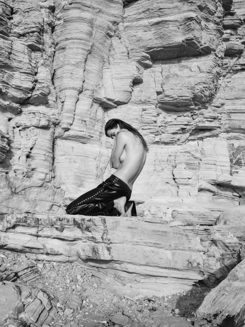 Monochrome shot of an adult female kneeling on a rock formation, posing with arms crossed covering her chest in a desert landscape.
