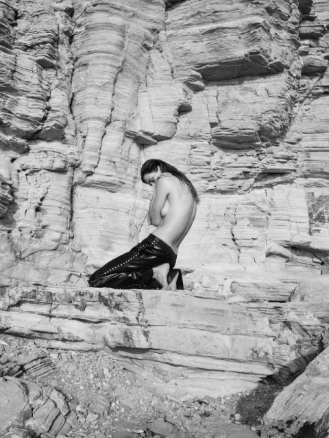 Monochrome shot of an adult female kneeling on a rock formation, posing with arms crossed covering her chest in a desert landscape.