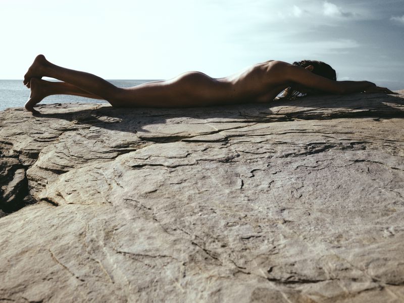 Adult female lying nude on a rock formation near the ocean with a clear sky in the background. Day light, outdoor setting.