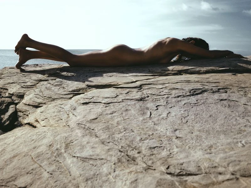 Adult female lying nude on a rock formation near the ocean with a clear sky in the background. Day light, outdoor setting.