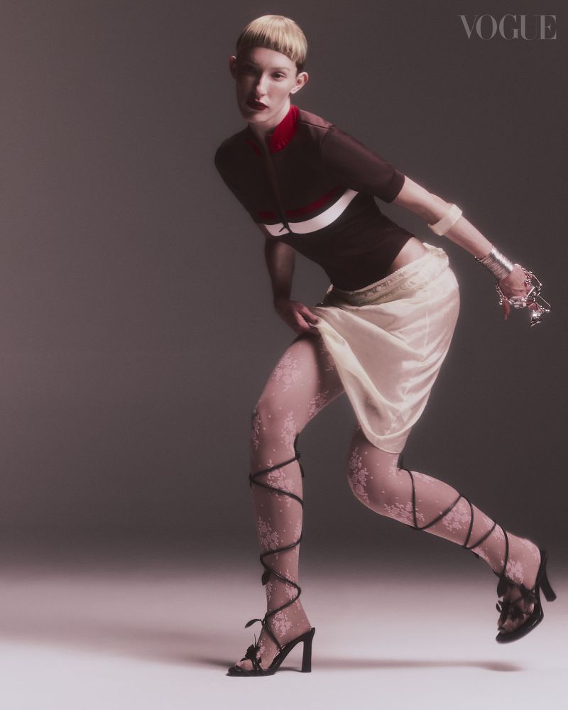A Vogue fashion editorial featuring a light-skinned woman in a brown top, cream skirt, decorative floral tights, and black heels posing in studio.