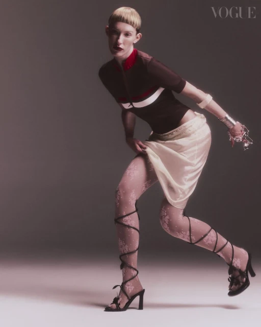 A Vogue fashion editorial featuring a light-skinned woman in a brown top, cream skirt, decorative floral tights, and black heels posing in studio.