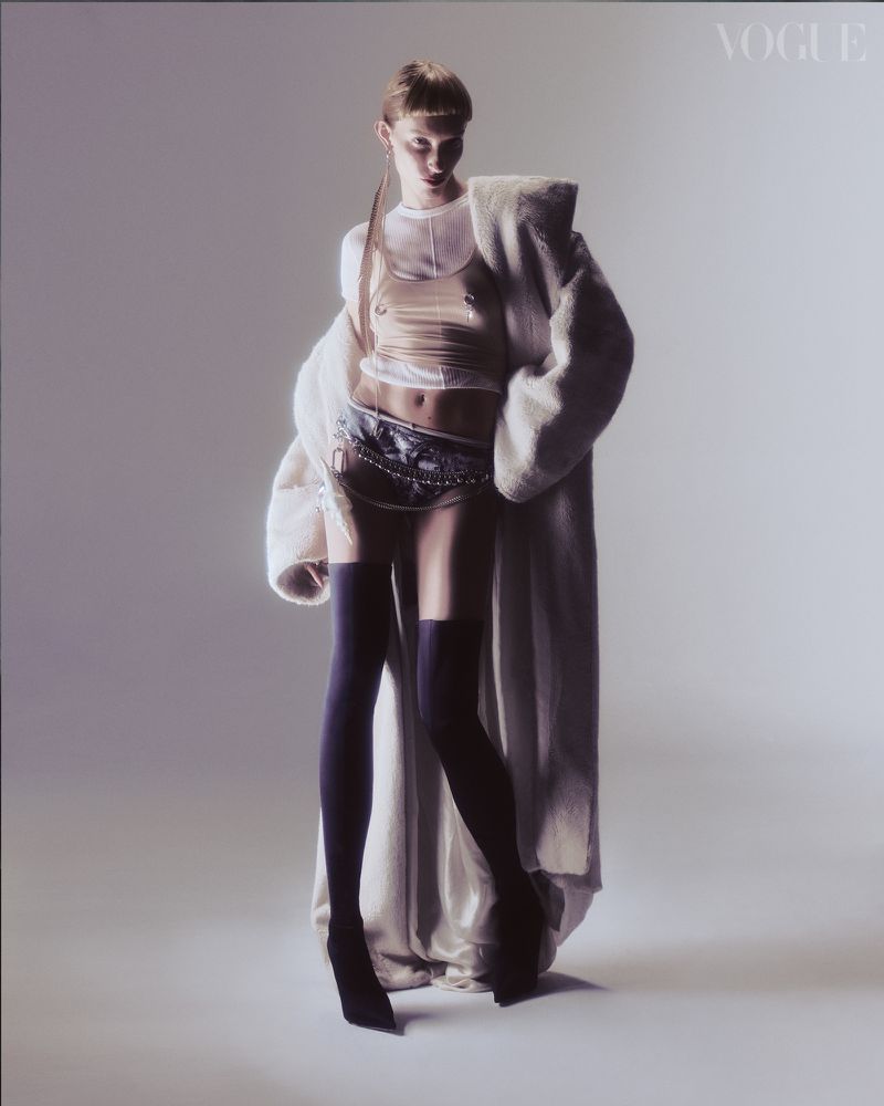 A tall, slim fashion model with translucent top and black stockings poses in a studio with a long, light coat over her shoulder.