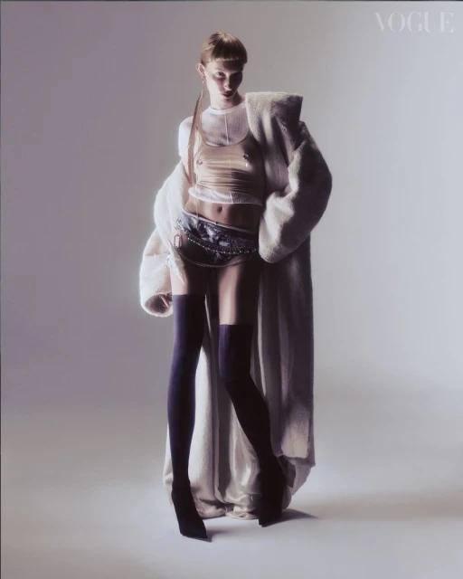 A tall, slim fashion model with translucent top and black stockings poses in a studio with a long, light coat over her shoulder.