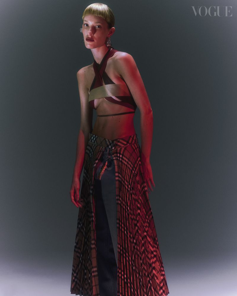 A model posing in a studio with a halter top and plaid skirt, under red and white lighting, evokes a fashion-forward editorial.