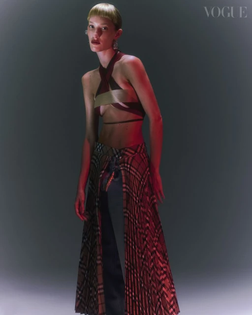 A model posing in a studio with a halter top and plaid skirt, under red and white lighting, evokes a fashion-forward editorial.