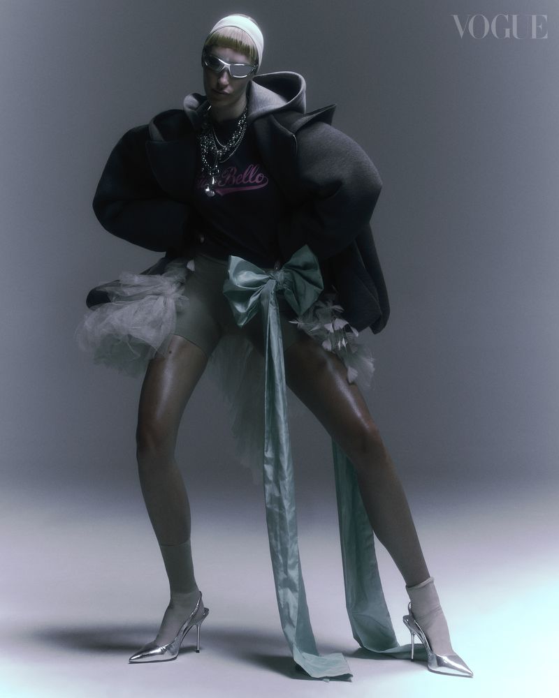 A fashion model poses in an oversized hoodie, tulle skirt, and silver shoes in a studio setting with soft lighting. Vogue logo visible.