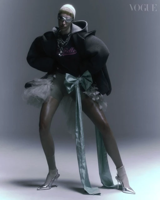 A fashion model poses in an oversized hoodie, tulle skirt, and silver shoes in a studio setting with soft lighting. Vogue logo visible.