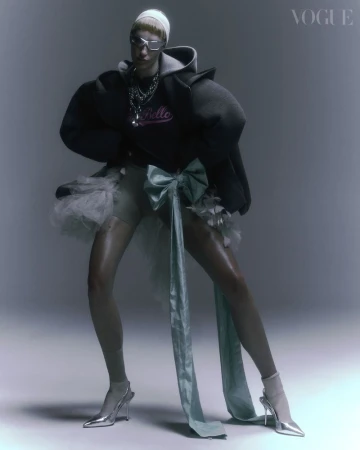 A fashion model poses in an oversized hoodie, tulle skirt, and silver shoes in a studio setting with soft lighting. Vogue logo visible.